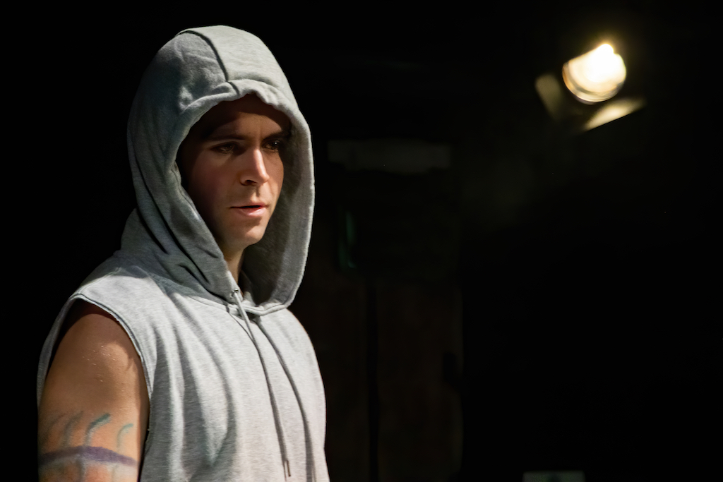 Actor wears a sleeveless hoodie and looks toward the camera with a stage light over his left shoulder